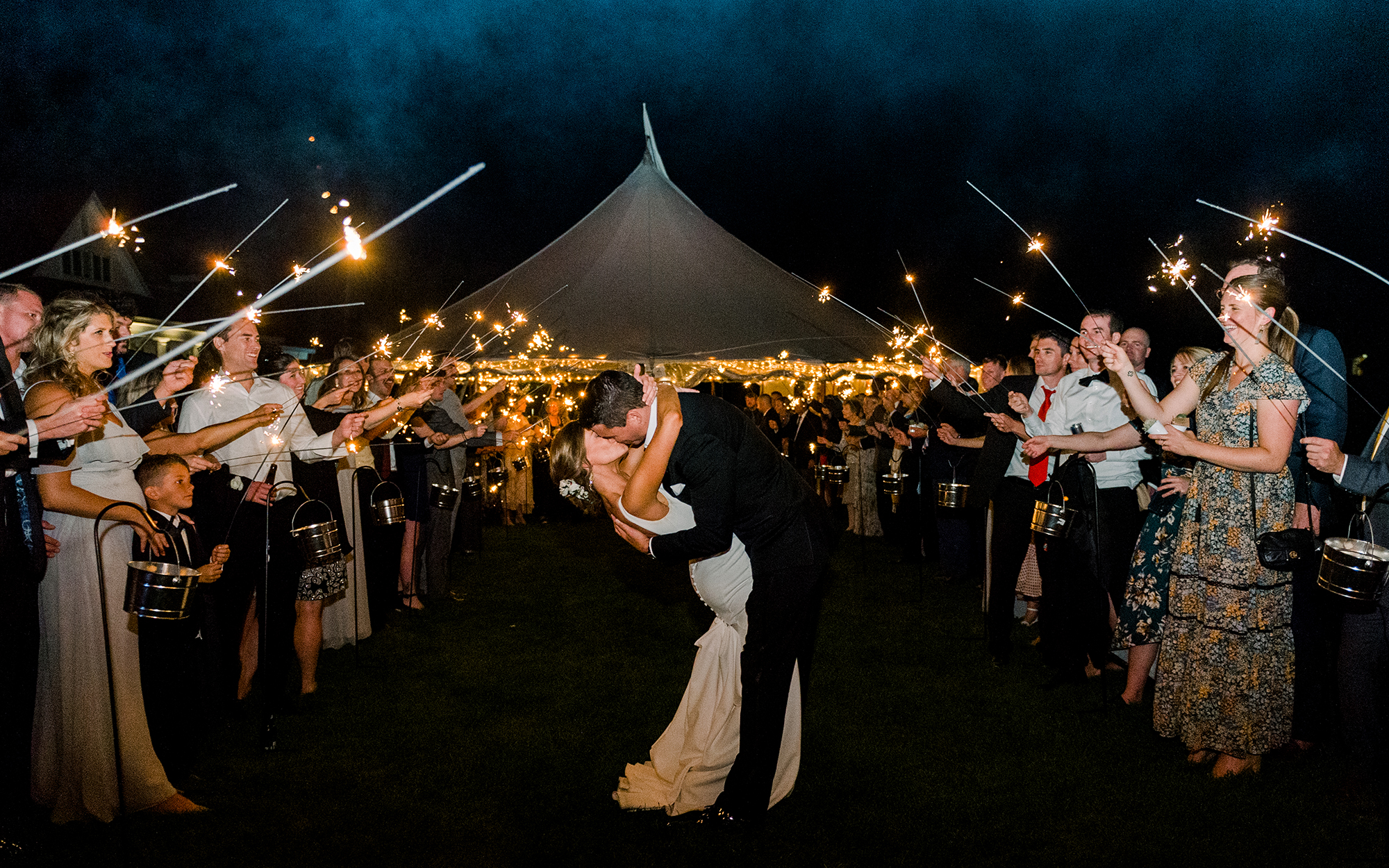 Bride and groom kissing surrounded by wedding party outdoors at night