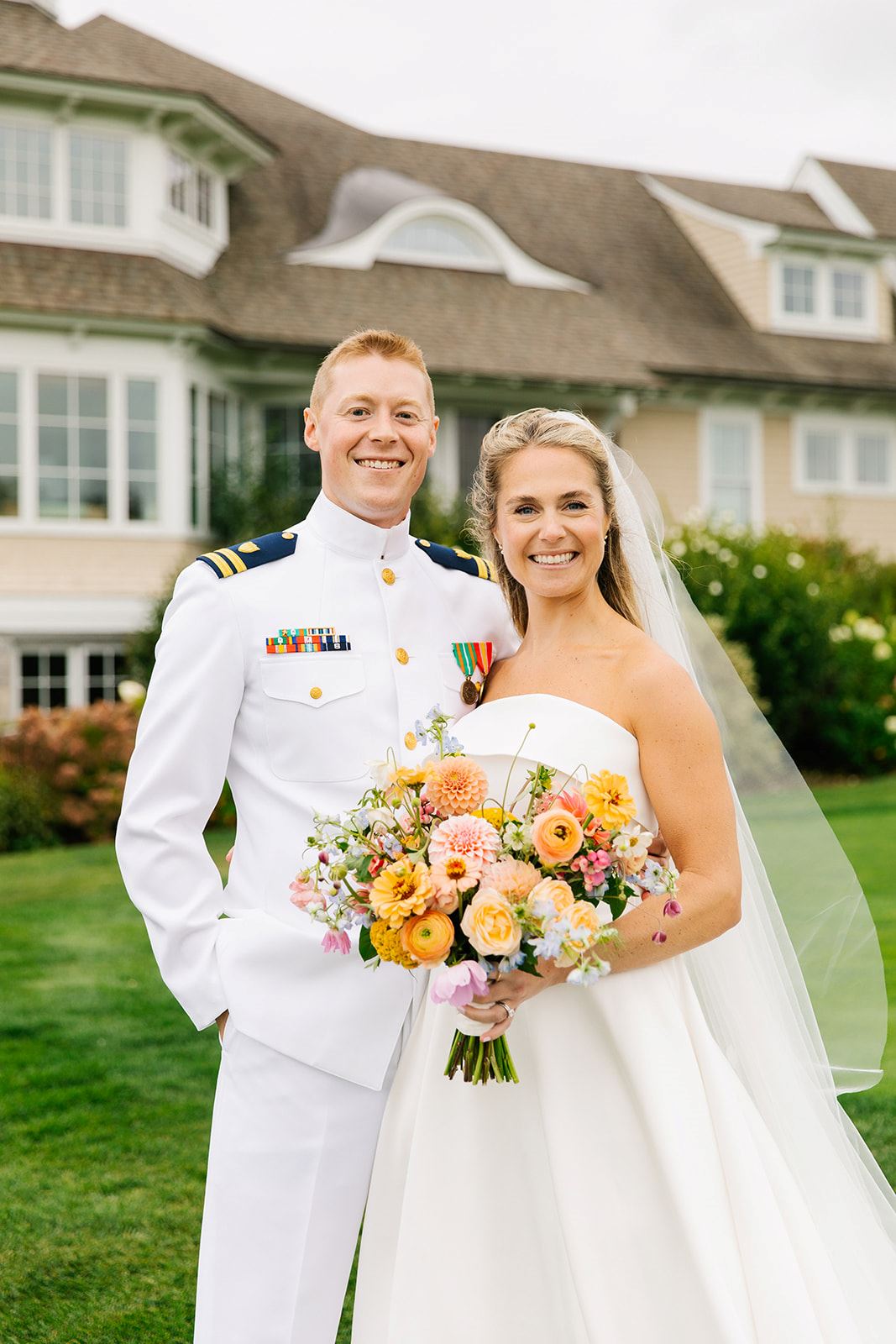 A bride and groom in military uniforms pose together in front of a grand house, showcasing their love and commitment.