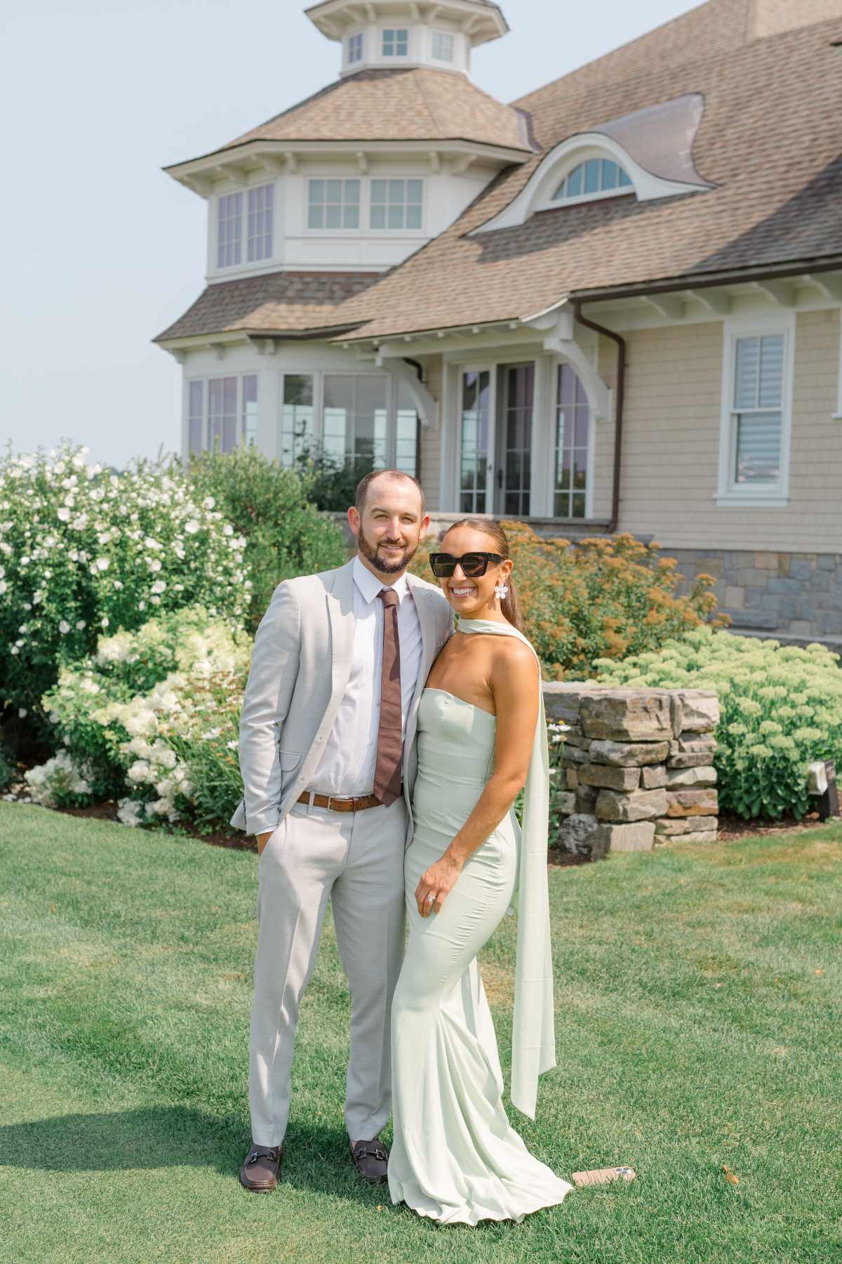 A man and woman in formal attire pose elegantly in front of a luxurious house, celebrating their wedding day.