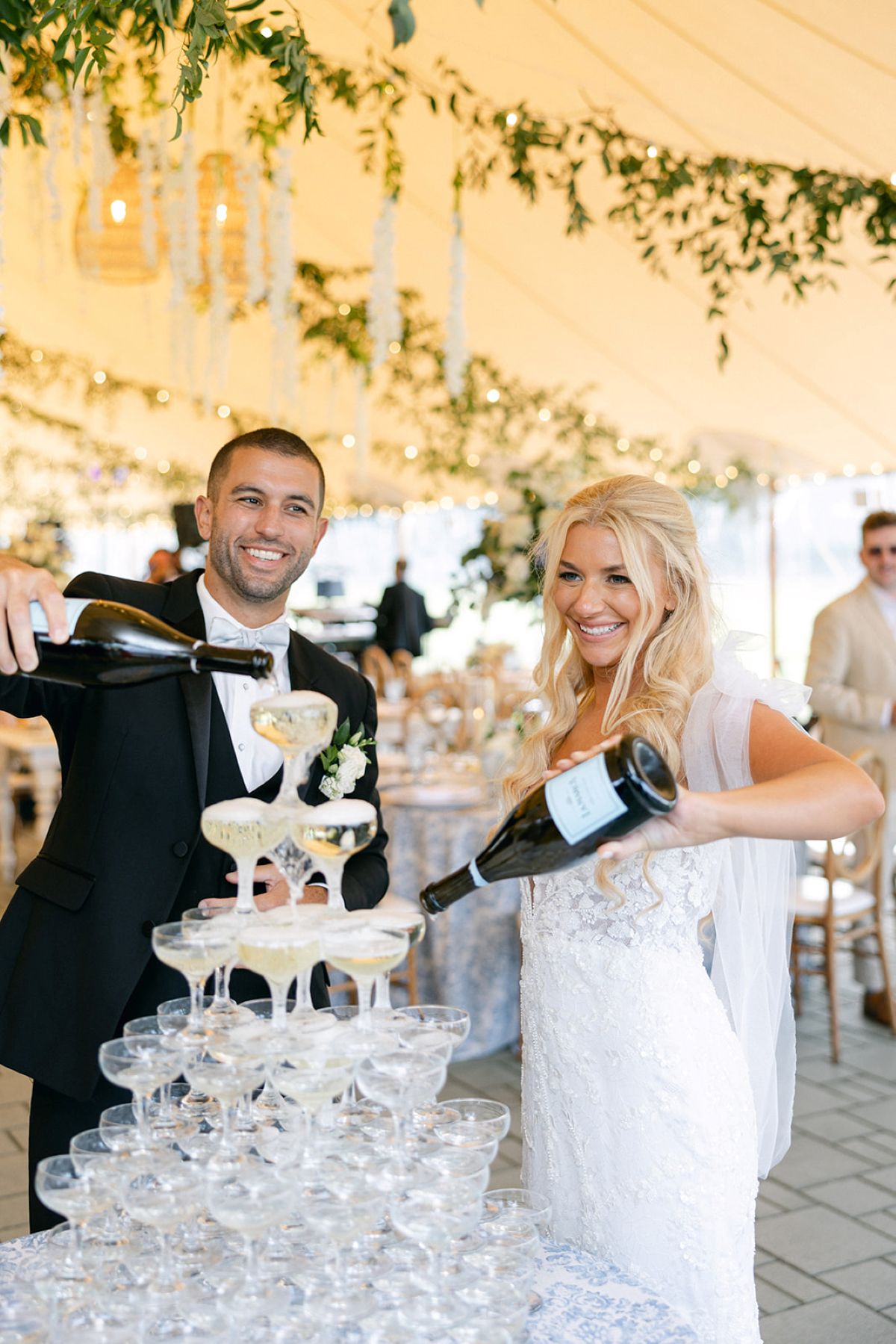 A bride and groom elegantly pour champagne into a towering glass structure at a luxury wedding celebration.