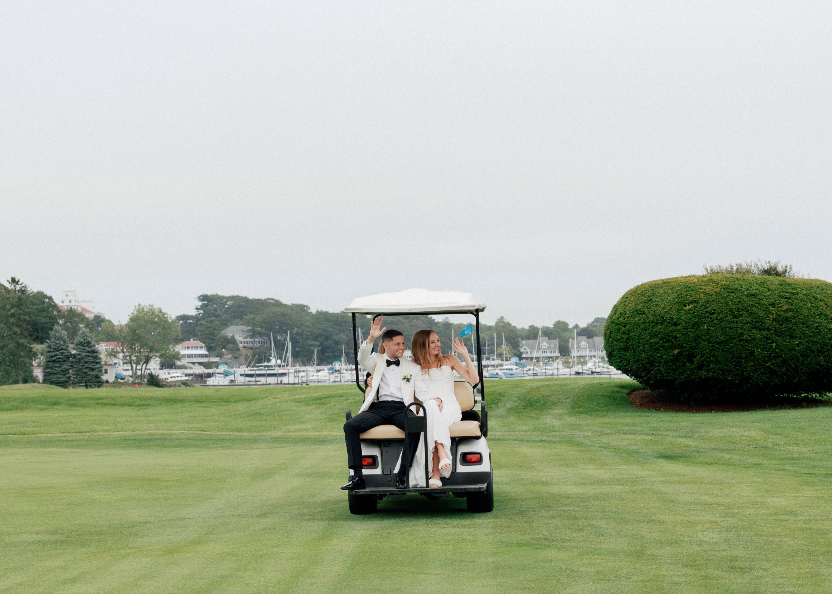 Newlywed couple dressed in wedding attire waving while riding a golf cart on a green lawn with a marina and houses in the background